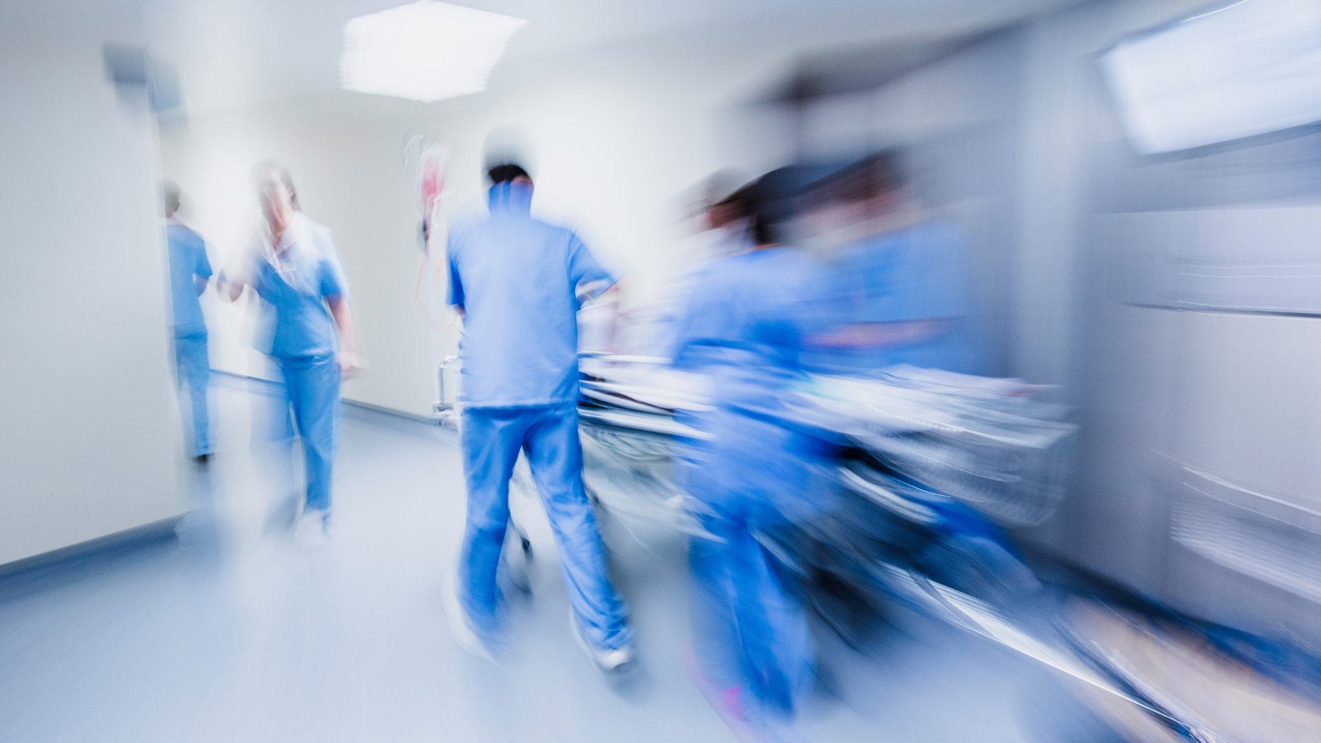 A blurry photo of multiple medical professionals wearing blue scrubs, rushing through a hospital corridor.