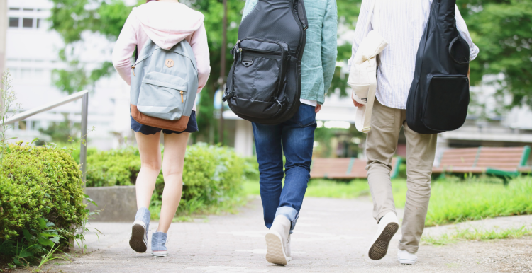 Three people walking away along a paved path in a green outdoor setting, each carrying backpacks; two have guitar cases on their backs, suggesting students walking on a campus.