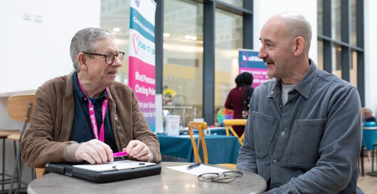 Two men sitting at a table, talking to each other. One man is wearing a Healthwatch lanyard.