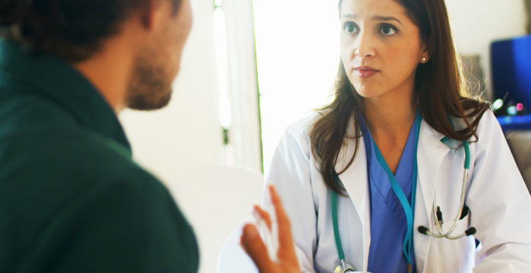 A man talking to a female doctor in a clinical setting. The woman is wearing a white medical coat with a stethoscope.