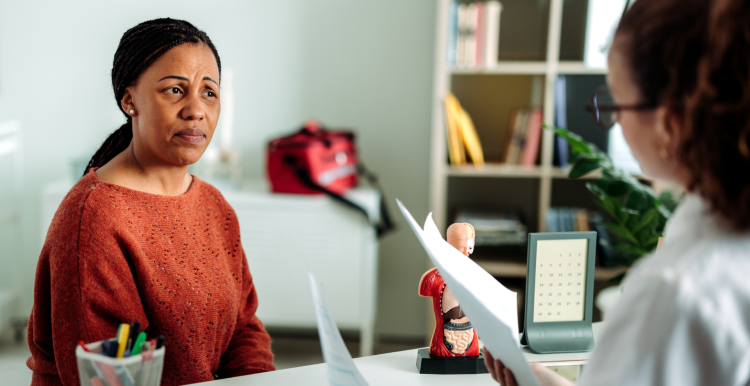 A woman discussing health concerns with female doctor. They are both sitting at the doctor's desk and the doctor is holding several sheets of paper.