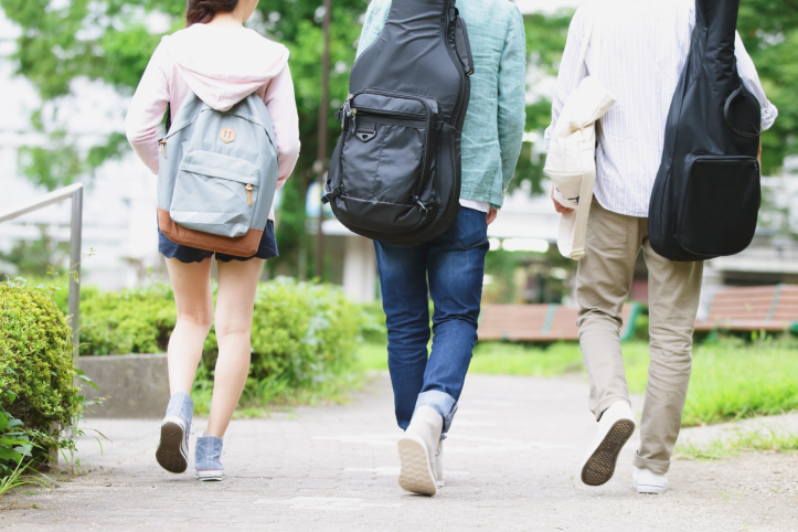Three people walking away along a paved path in a green outdoor setting, each carrying backpacks; two have guitar cases on their backs, suggesting students walking on a campus.