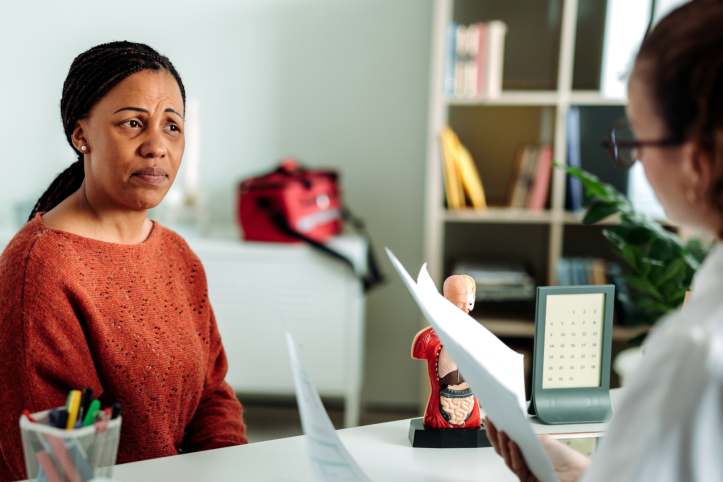 A woman discussing health concerns with female doctor. They are both sitting at the doctor's desk and the doctor is holding several sheets of paper.