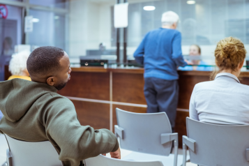 Rear view of people sitting in a hospital waiting room. One person is checking in at a main desk.