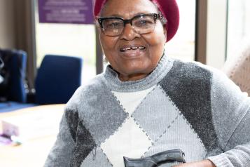 An older black woman smiling. She is wearing a grey jumper and red berets. She is holding a walking stick.