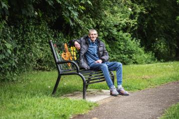 A man sitting on a park bench, with trees and bushes in the background.