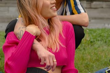 A trans couple sit on a blanket in the park, smiling and sharing a moment together. One person with long pink hair, a bright pink top, and colorful trousers reclines on the blanket, smiling up at their partner. The other person, wearing a striped shirt, leans over them with an affectionate gaze.