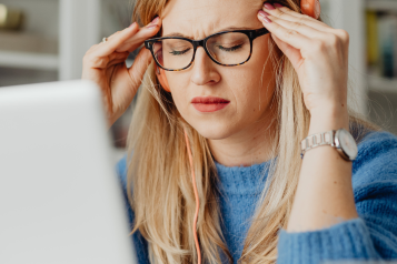A woman sitting in front of a laptop. She is wearing headphones and holding her head as if she has a headache.