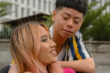 A trans couple sit on a blanket in the park, smiling and sharing a moment together. One person with long pink hair, a bright pink top, and colourful trousers reclines on the blanket, smiling up at their partner. The other person, wearing a striped shirt, leans over them with an affectionate gaze.