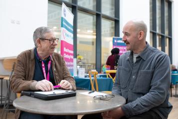 Two men sitting at a table, talking to each other. One man is wearing a Healthwatch lanyard.