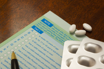 A green prescription form on a wooden surface. On top of the form is a blister pack of tablets, some loose tablets, and a pen.