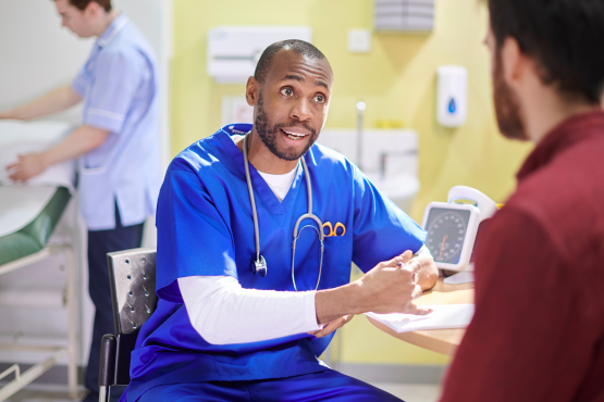 A medical professional wearing blue scrubs, talking to a patient at a table in a hospital. Behind them, a second medical professional is making up a hospital bed.