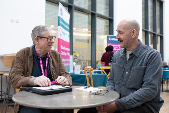 Two men sitting at a table, talking to each other. One man is wearing a Healthwatch lanyard.