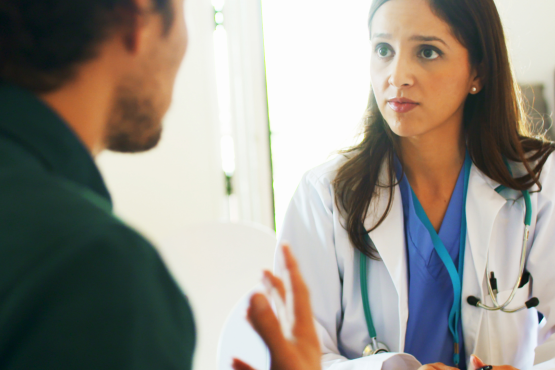 A man talking to a female doctor in a clinical setting. The woman is wearing a white medical coat with a stethoscope.