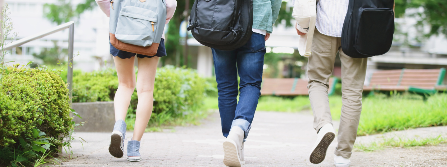 Three people walking away along a paved path in a green outdoor setting, each carrying backpacks; two have guitar cases on their backs, suggesting students walking on a campus.