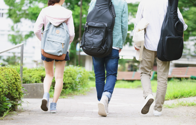 Three people walking away along a paved path in a green outdoor setting, each carrying backpacks; two have guitar cases on their backs, suggesting students walking on a campus.