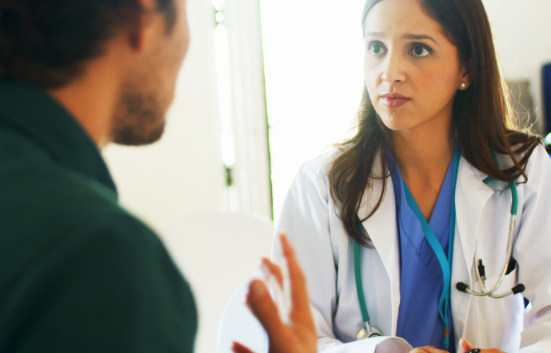 A man talking to a female doctor in a clinical setting. The woman is wearing a white medical coat with a stethoscope.