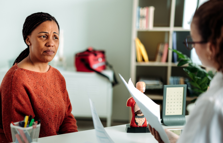 A woman discussing health concerns with female doctor. They are both sitting at the doctor's desk and the doctor is holding several sheets of paper.