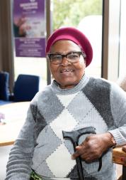 An older black woman smiling. She is wearing a grey jumper and red berets. She is holding a walking stick.