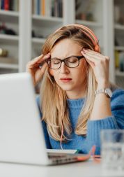 A woman sitting in front of a laptop. She is wearing headphones and holding her head as if she has a headache.
